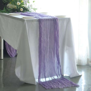 May include: A white tablecloth is draped with a lavender table runner. The runner is made of a sheer, crinkled fabric. White plates and a floral arrangement are on the table. The table is set on a gray floor.