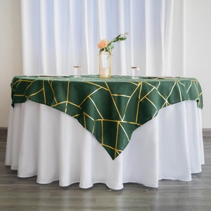May include: A green square tablecloth with a gold geometric pattern. The tablecloth is placed on a white round tablecloth.