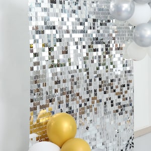 May include: A silver sequin backdrop with a reflective surface, perfect for parties and events. The backdrop is adorned with square sequins, creating a shimmering effect. Gold, white, and silver balloons are in the foreground.