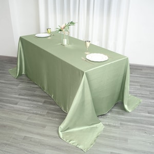 May include: A sage green satin tablecloth draped over a rectangular table. The tablecloth has a smooth, silky texture and hangs down on all sides of the table. There are two white plates, two wine glasses, and a vase with flowers on the table.