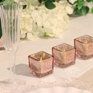 May include: Three square glass candle holders with a rose gold finish. The candle holders are sitting on a white tablecloth.