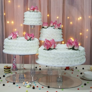 May include: Four tiered clear acrylic cake stand with four white cakes decorated with pink rose accents. The cakes are on a white tablecloth with colorful sprinkles.