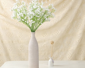 May include: A tall, textured, off-white vase holds a bouquet of white baby's breath flowers. A small white bottle with reeds and a decorative ball sits to the right. A tan ribbon and black scissors are on the table.