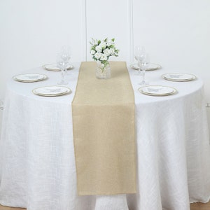 May include: A beige burlap table runner on a white tablecloth with white plates and glasses. A vase with white flowers sits in the center of the runner.