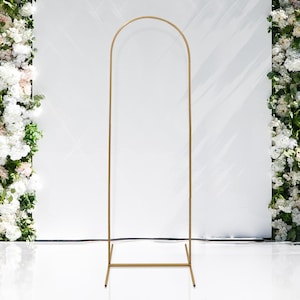 May include: A tall, gold metal archway, ideal for weddings or events. The arch has a rounded top and a rectangular base for stability. The background is a white wall with floral arrangements on either side.