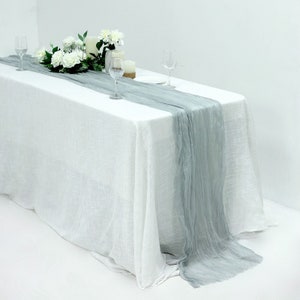 May include: A white tablecloth with a light gray runner down the center. The runner is made of a sheer fabric and has a slightly wrinkled texture. There are two wine glasses and a bouquet of white flowers on the table.