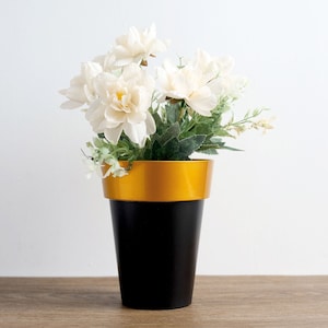 May include: A black and gold planter with white flowers. The planter has a gold rim and a black base.