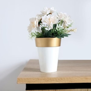 May include: White ceramic planter with a gold rim, filled with a bouquet of white flowers and green foliage.