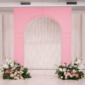 May include: A pink archway backdrop for events, with a textured surface and an arched opening. Two floral arrangements with white, pink, and green flowers are placed on either side of the archway, creating a decorative setting for weddings or parties.
