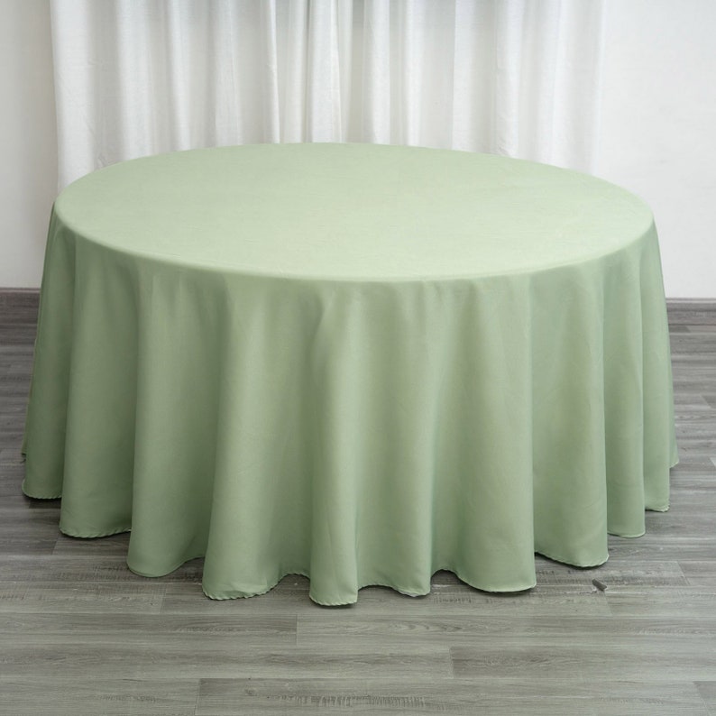 Op de afbeelding: Een ronde, saliegroene tafelkleed met een gladde, kreukvrije afwerking. Het tafelkleed is over een tafel gelegd en hangt tot op de grond.