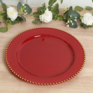 May include: A round, red charger plate with a gold beaded rim. The plate is made of a glossy material and is suitable for decorative purposes. The plate is approximately 12 inches in diameter.