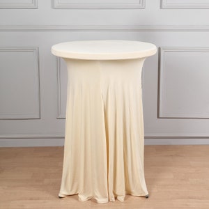 May include: A round, cream-colored table with a fitted, floor-length cover. The table has a simple, elegant design, suitable for events or gatherings. The table's height is approximately 42 inches.