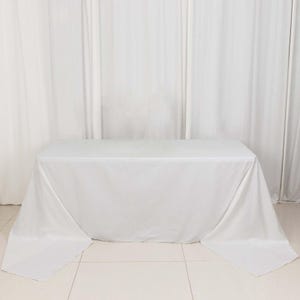 May include: A white rectangular tablecloth draped over a table. The fabric is smooth and appears to be made of a lightweight material. The table is centered in front of white curtains.