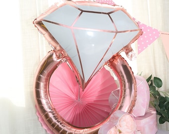 Könnte beinhalten: Ein großer roségoldener Diamantring-Ballon mit einem weißen rautenförmigen Mittelteil. Der Ring ist vor einem rosa Papierfächer platziert und von rosa und weißen Dekorationen umgeben, ideal für eine Brautparty oder Verlobungsfeier.