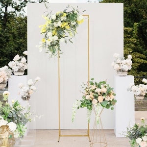 May include: A gold metal archway backdrop with white flowers and greenery. The archway is standing on a white surface. There are other floral arrangements on white pedestals.