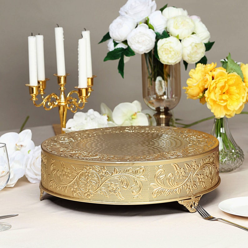 May include: Gold metal cake stand with an ornate floral design. The stand has three legs and is round. It is on a white tablecloth with white flowers and candles in the background.