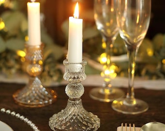 May include: Two clear glass candle holders with a diamond pattern. Each holder has a white pillar candle. The candles are lit and the flames are visible.