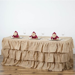 May include: A three-tiered burlap table skirt with a ruffled edge. The skirt is beige and has a natural, rustic look. It is shown draped over a rectangular table with white plates, wine glasses, and burgundy napkins.