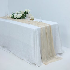 May include: A white tablecloth with a beige sheer table runner. The table is set with two glasses and a plate. There are white flowers and candles on the table.