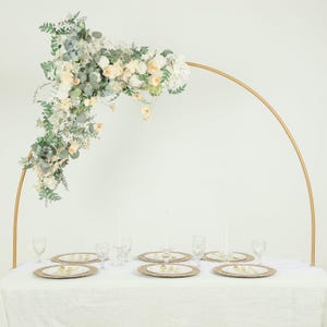 May include: A wedding reception table setting with a gold arch decorated with white and cream-colored flowers. The table is set with gold-rimmed plates, silverware, and clear glasses. White candles are lit, creating an elegant and romantic atmosphere.