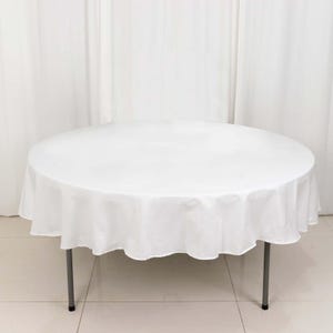 May include: A round white tablecloth covers a table with gray metal legs. The tablecloth drapes over the edge of the table. The background is a white curtain.