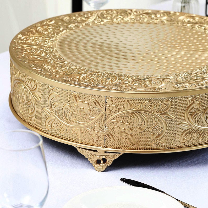 May include: Gold cake stand with an ornate floral design and three legs. The stand is round and has a textured surface.