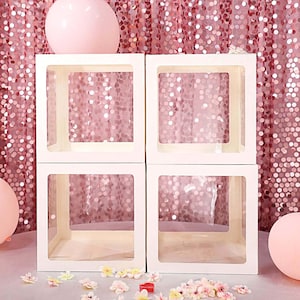 May include: Four white square boxes with clear sides, stacked two high. The boxes are in front of a pink sparkly background.