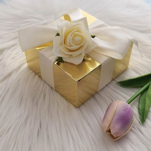 May include: A gold gift box with a white satin ribbon and a white rose on top. A single purple tulip sits on a white fluffy surface next to the box.