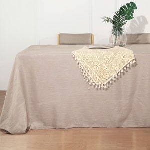 May include: A beige linen tablecloth with a white crocheted table runner with tassels. The table runner has a delicate floral pattern.