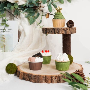 May include: A rustic, two-tiered wooden cake stand displays several cupcakes. One cupcake is decorated to resemble a Christmas tree with a gold star. Other cupcakes have white frosting and red berry accents. A pine cone and greenery complete the display.
