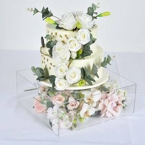 May include: A two-tiered white wedding cake decorated with gold accents and white flowers, displayed on a clear acrylic cake stand. The cake is surrounded by white and pink flowers.
