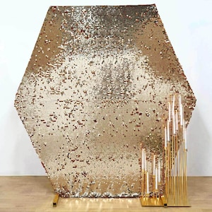 May include: A large, hexagonal gold sequin backdrop with a matching gold stand. In front of the backdrop are several gold candle holders with white candles. The backdrop and candle holders are for event decoration.