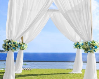 May include: White sheer fabric drapes are hung from a wooden archway, creating a beautiful backdrop for an outdoor wedding ceremony. The archway is decorated with white flowers and blue accents, and the ocean is visible in the background.
