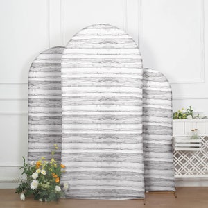 May include: Three white wooden arch backdrops with a gray woodgrain pattern. The backdrops are different sizes and are standing on a wooden floor.