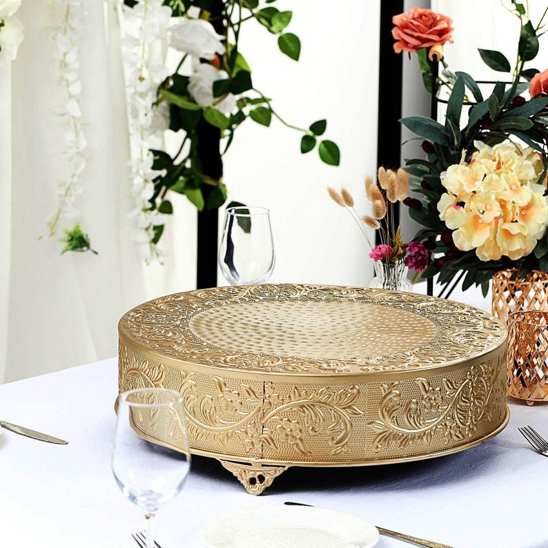 May include: Gold metal cake stand with an ornate design. The stand has three legs and a round top. The stand is on a white tablecloth.