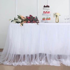 May include: A white tulle table skirt with a ruffled edge. The table is set with a tiered cupcake stand, floral arrangements, and a gold vase. Cupcakes in various colors are displayed on the stand.