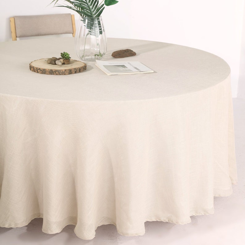 May include: A round, off-white tablecloth with a subtle texture. The tablecloth is draped over a round table and has a slight ruffle around the edge.