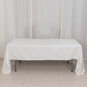 May include: A white rectangular tablecloth draped over a folding table with silver legs. The table is set against a white curtain backdrop. The tablecloth appears to be made of a smooth, wrinkle-resistant fabric.
