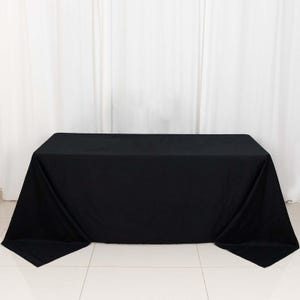 May include: A black rectangular tablecloth draped over a table. The tablecloth is made of a solid black fabric and covers the entire table, with the corners hanging down. The background is a white curtain.
