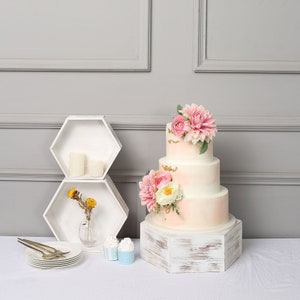 May include: A four-tiered wedding cake with pink and white frosting, adorned with pink and white flowers and gold accents. The cake sits on a white, distressed wooden stand. Two white hexagon shelves, candles, and cupcakes are also visible.