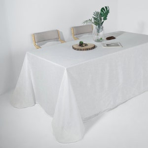 May include: A white rectangular tablecloth covers a table, with a slight texture visible. Two chairs with light gray seats and wooden frames are in the background. A clear vase with green leaves and a small wooden slice with decorative items are on the table.