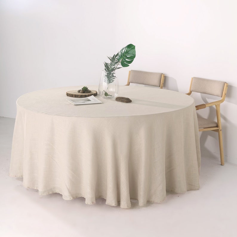 May include: A round, off-white linen tablecloth draped over a table with two chairs. The tablecloth has a slight sheen and is wrinkle-free. A small potted plant and a book are on the table.