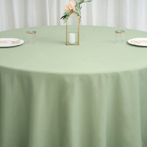 Op de afbeelding: Een ronde, lichtgroene tafelkleed met een kleine gouden decoratieve standaard met een enkele witte kaars en een klein boeket roze bloemen. Twee heldere glazen bekers staan op de tafel.