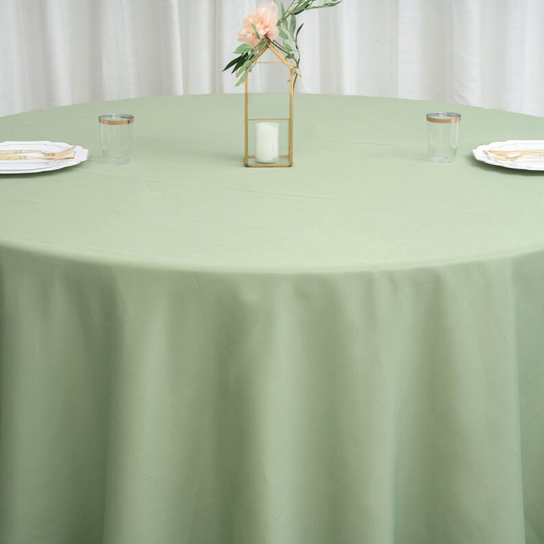Op de afbeelding: Een ronde, lichtgroene tafelkleed met een kleine gouden decoratieve standaard met een enkele witte kaars en een klein boeket roze bloemen. Twee heldere glazen bekers staan op de tafel.
