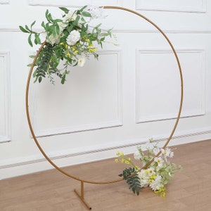 May include: A gold metal circular wedding arch decorated with white and yellow flowers and green foliage. The arch is supported by a base and is set against a white wall.