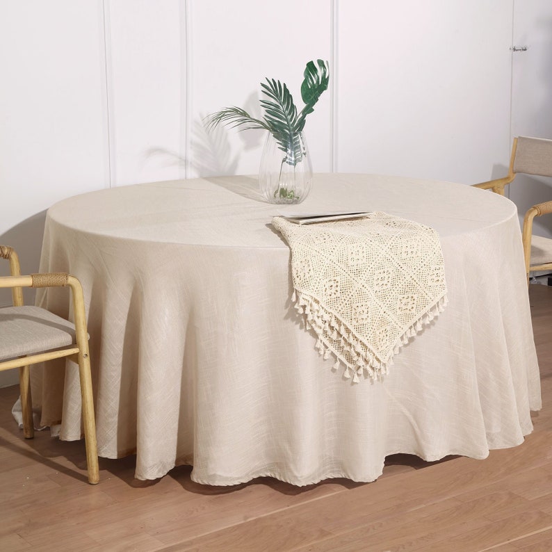 May include: A round, beige tablecloth with a crocheted table runner with fringe. The tablecloth is on a round table with a vase of green leaves in the center.