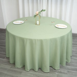 Op de afbeelding: Een ronde, saliegroene tafelkleed met een gegolfde rand. Het tafelkleed ligt op een houten vloer en heeft een klein bloemstuk in het midden.