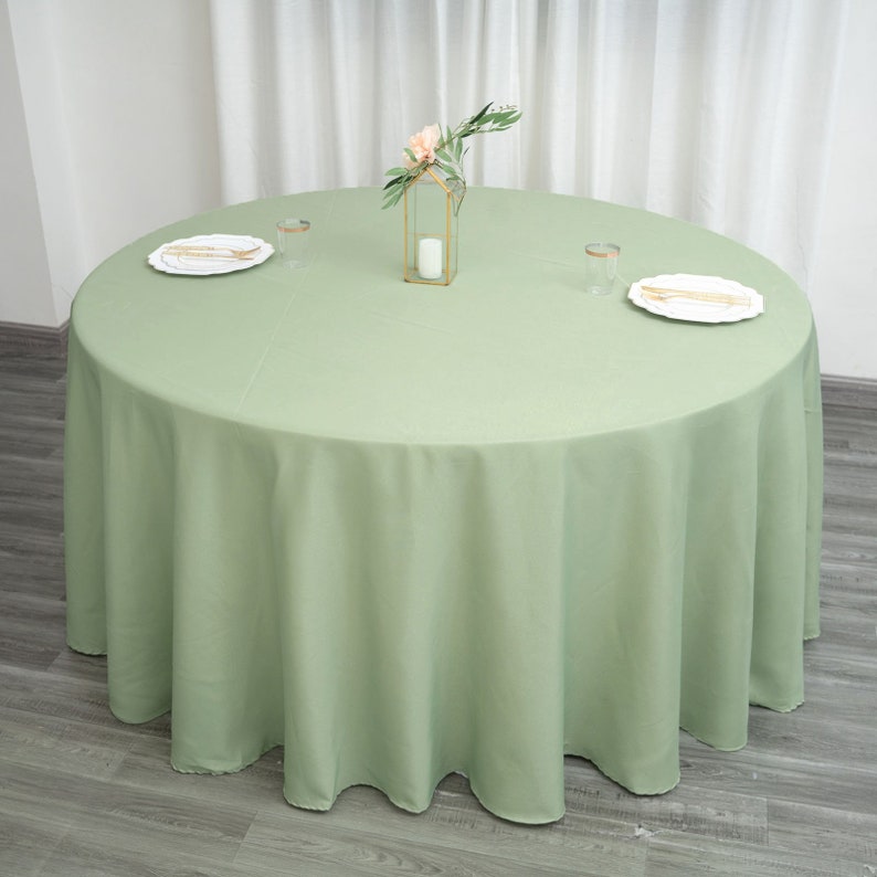 Op de afbeelding: Een ronde, saliegroene tafelkleed met een gegolfde rand. Het tafelkleed ligt op een houten vloer en heeft een klein bloemstuk in het midden.