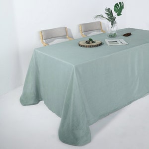 May include: A light green rectangular tablecloth draped over a table. The tablecloth is made of a textured fabric. Two chairs with light gray seats and wooden frames are in the background. A vase with green leaves and a wooden coaster are on the table.