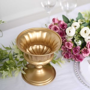 May include: A gold-colored metal urn-shaped vase with a wide, fluted top and a pedestal base. The vase is empty and sits on a white tablecloth, with flowers and greenery in the background. The vase is approximately 7 inches tall.
